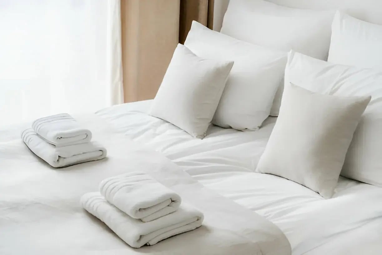 A neatly made bed with white linens, multiple white and cream-colored pillows, and two sets of folded white towels resting at the foot of the bed.