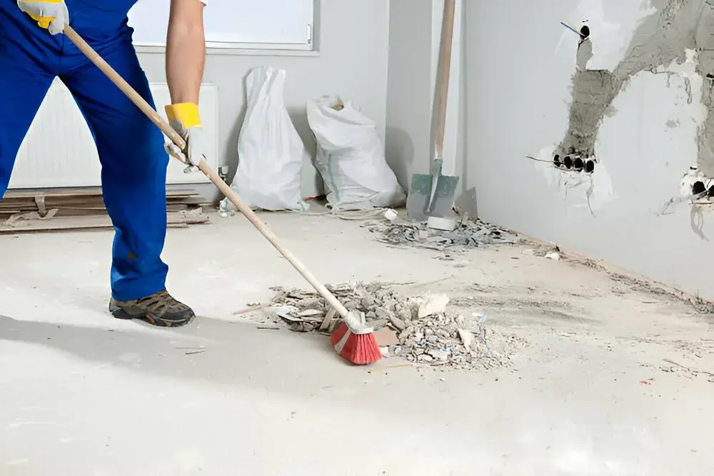 A construction worker in blue overalls and work gloves using a red-bristled broom