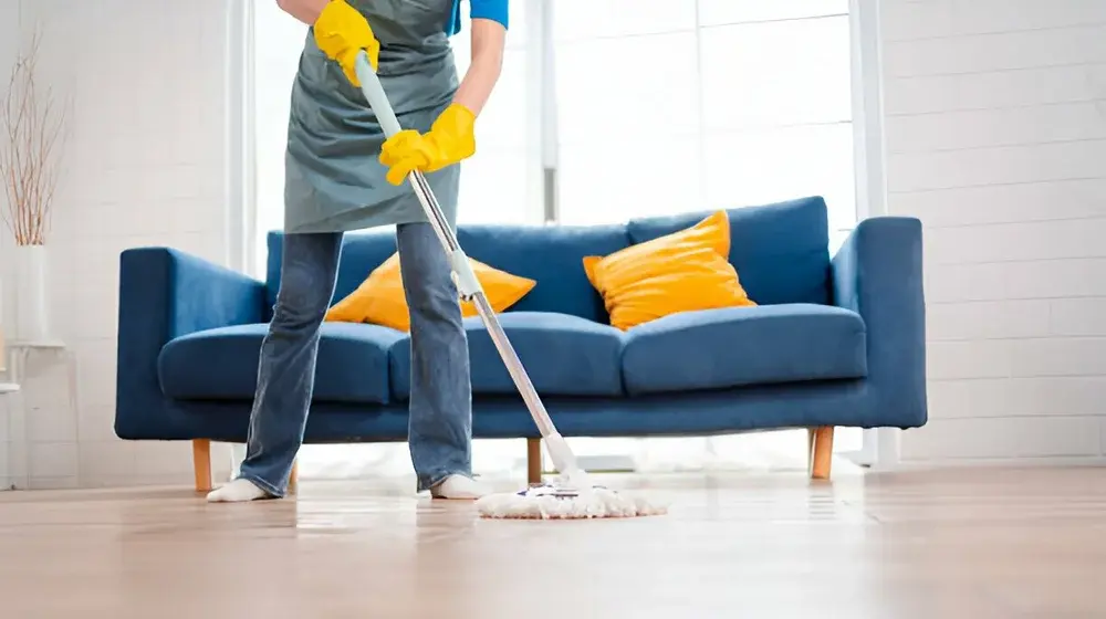 A person wearing yellow gloves and an apron is mopping a light-colored wood floor in a living room with a blue sofa and yellow accent pillows.